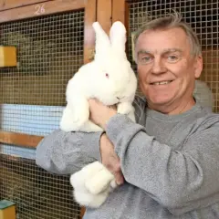 Ein Mann hält ein weißes Kaninchen mit roten Augen. Hinter ihnen sind Käfige mit Nummern.