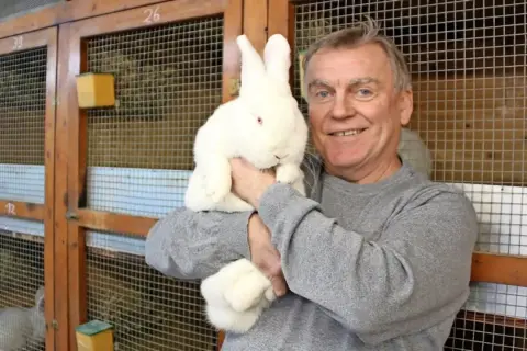 Ein Mann hält ein weißes Kaninchen mit roten Augen. Hinter ihnen sind Käfige mit Nummern.